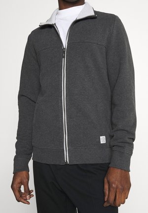 Mężczyzna w ciemnoszarej kurtce na suwak na białej koszuli i czarnych spodniach, stojący na jednolitym białym tle.