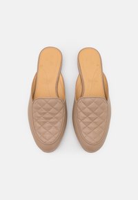 Sapatos slip-on de couro bege com um padrão acolchoado na parte superior, apresentando uma biqueira arredondada e laterais abertas para facilitar o uso.