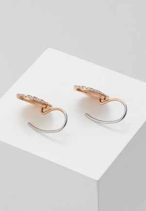 Paar goldene und silberne Creolen mit eingearbeiteten kleinen klaren Edelsteinen, präsentiert auf einem weißen Würfel vor grauem Hintergrund.