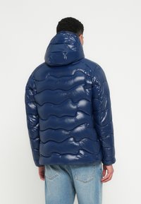 Jachetă puffer albastru marin, cu o textură lucioasă și matlasată, cu glugă atașată, având linii de design curbe și manșete elastice.