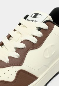 Λευκό και καφέ δερμάτινο αθλητικό παπούτσι με υφασμένα πάνελ, επίπεδα κορδόνια, λογότυπο και σκούρα λατέξ σόλα. Διαθέτει διατρήσεις στην μύτη για αναπνευστικότητα.