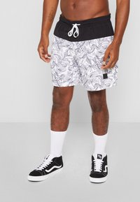 Svømmebukser med sort talje og hvid bund, med et sort og hvidt bladmønster. Kombineret med sorte sneakers og hvide strømper.