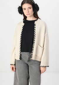 Beige gestrickte Strickjacke mit schwarzem Kontrastbesatz entlang der vorderen Kanten und an den Bündchen; verfügt über eine Tasche auf der linken Seite. Getragen über einem schwarzen Oberteil.
