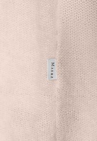 Ljust rosa stickad tyg med en strukturerad yta. En liten vit etikett med "MAERZ" är fäst vid tyget.