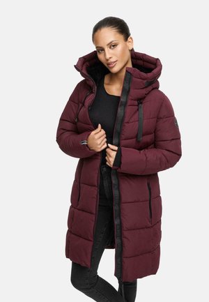 Vrouw in een lange, gevoerde bordeauxrode winterjas met capuchon, zwarte rits en zwarte broek tegen een effen lichte achtergrond.