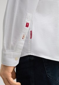 Camicia bianca a maniche lunghe con polsini abbottonati, caratterizzata da dettagli ricamati rossi e etichette sulla manica. Tessuto in cotone liscio.