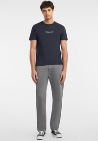 Jūros mėlynos medvilninės marškinėliai su baltu "GUESS JEANS" logotipu, derinami su šviesiai pilkomis kelnėmis ir juodais sportiniais bateliais su baltais padais.