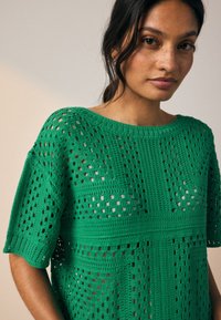 Roheline heegeldatud top, millel on lõdvestunud lõige, lühikesed varrukad ja ruudukujuline muster tekstuursete segmentidega.