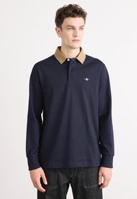 Polo a maniche lunghe blu navy con colletto in corduroy color tan, caratterizzato da un logo discreto sul petto e polsini a costine. Vestibilità casual e tessuto testurizzato.