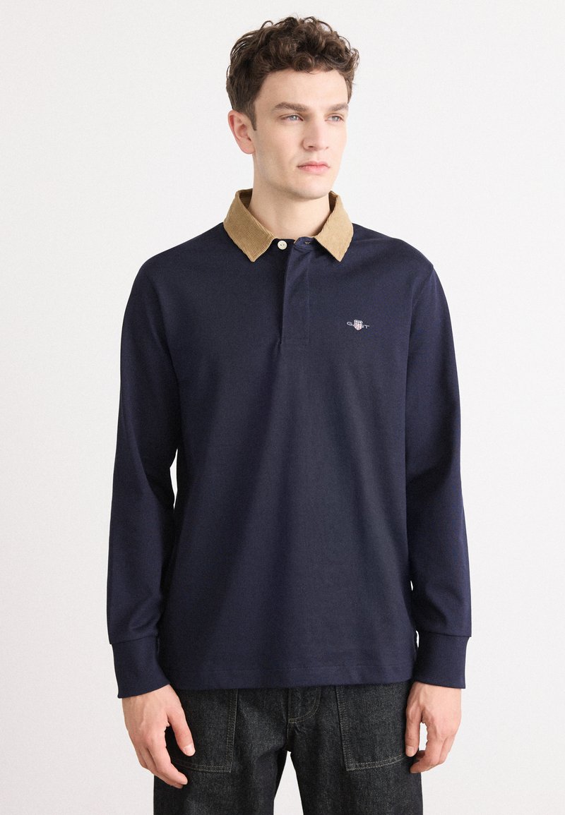 Polo a maniche lunghe blu navy con colletto in corduroy color tan, caratterizzato da un logo discreto sul petto e polsini a costine. Vestibilità casual e tessuto testurizzato.