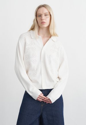 Szőke nő, aki túlméretezett, fehér, gombos kardigánt visel finom kivágásmintákkal és sötétkék, széles szárú farmernadrágot, kezét összekulcsolva áll.