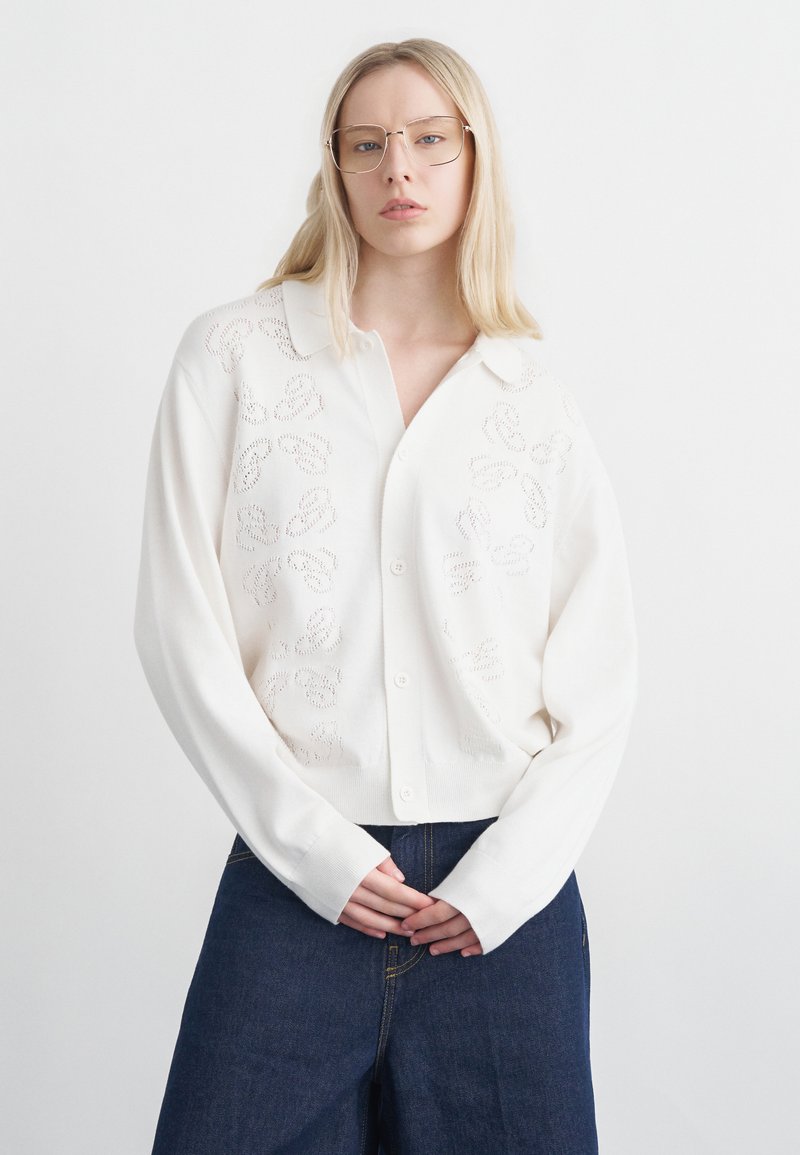 Ξανθιά γυναίκα που φοράει λευκό, oversized πουλόβερ με κουμπιά και διακριτικά μοτίβα κοψίματος, καθώς και σκούρο μπλε τζιν με φαρδιά μπατζάκια, στέκεται με τα χέρια σταυρωμένα.