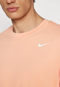 Camiseta Nike color durazno con cuello redondo, mangas cortas y un logo swoosh blanco en el pecho. Tela de algodón suave, diseño minimalista.