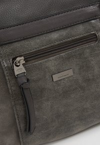 Sac en cuir gris avec une surface texturée, une poche zippée à l'avant et une plaque logo en métal. Le contour lisse contraste avec le corps mat.