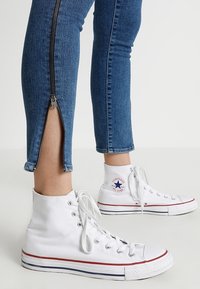 Bílé tenisky s vysokým střihem, zaoblenou špičkou, šněrováním a červenými a modrými pruhy na podrážce, doplněné modrými džínami s kovovým zipem.