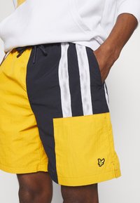 Costumi da bagno da uomo gialli e blu navy. Realizzati in tessuto leggero, presentano strisce in rete, una grande tasca laterale e un trucco con logo.