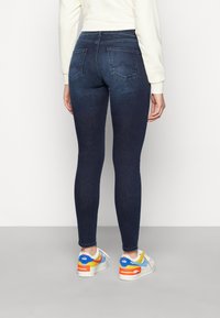 Persona che indossa jeans skinny blu scuro, una maglietta bianca a maniche lunghe e scarpe Nike Air bianche con accenti blu, arancione e giallo, in piedi di spalle.