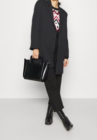 Schwarze strukturierte Handtasche mit strukturierter Oberfläche, kurzen Griffen und rechteckiger Form, kombiniert mit einem schwarzen Mantel und dunklen Hosen.