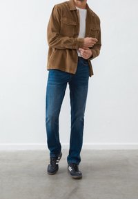 Brown corduroy shirt jacket, white t-shirt, blue jeans, and black sneakers with white stripes, displayed against a plain white background.