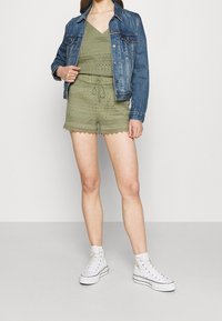 Olivově zelený top s háčkovaným vzorem a odpovídající kraťasy se zoubkovanými okraji, doplněný džínsovou bundou a bílými teniskami na vysoké podrážce.