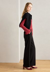 Red long-sleeve top under black sleeveless wrap, paired with black wide-leg trousers and burgundy block-heeled shoes on wooden floor.