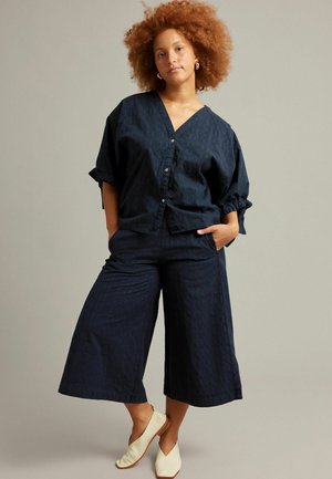 Vrouw met krullend haar, gekleed in een marineblauw, geknoopt blouse en wijde cropped broek, staand met handen in de zakken en witte instapschoenen.