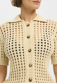 Cárdigan de punto color crema con cuello tipo solapa, que presenta un patrón de cuadrícula, mangas cortas y grandes botones marrones.