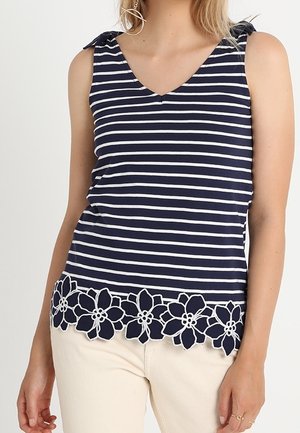 Nainen, joka pukeutuu hihattomaan, tummansinivalkoraidalliseen V-kaulaiseen toppiin, jossa on kukallinen pitsireuna, yhdistettynä vaaleanbeigeihin housuihin.