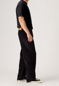 Homme portant un pantalon en velours côtelé noir, un t-shirt noir et des baskets noires, debout devant un fond clair uni, vue de profil.