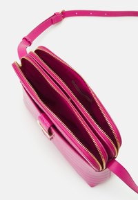Różowa torebka na ramię wykonana z teksturowanego materiału, wyposażona w trzy kieszenie na zamki z złotymi okuciami oraz smukły regulowany pasek.