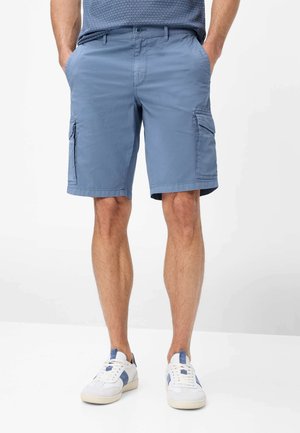 Mann trägt blaue Cargo-Shorts und weiße Sneaker mit blauen Akzenten und steht mit Händen in den Taschen vor einem einfarbigen Hintergrund.