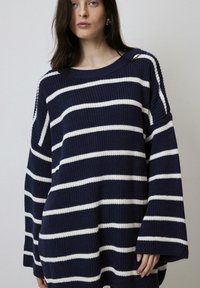 Pull en maille oversized bleu marine avec des rayures blanches horizontales, col large et épaules tombantes. Tissu texturé avec une coupe ample.