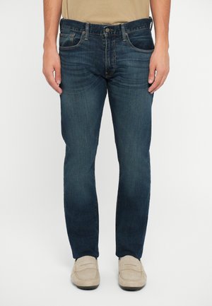 Jean en denim bleu foncé avec une coupe droite, cinq poches et des éléments en métal argenté, porté avec des mocassins beige clair.