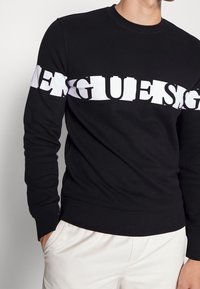 Homme portant un sweat-shirt noir à manches longues avec un texte blanc audacieux et déformé sur la poitrine et les bras, assorti à un pantalon blanc.