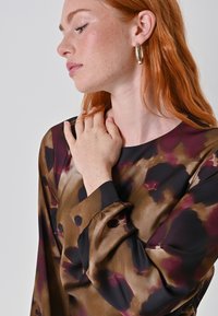 Bruine en zwarte blouse met een patroon van paarse en tan vlekken; heeft een ronde hals en gathered manchetten. Model raakt voorzichtig haar sleutelbeen aan.