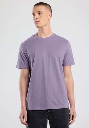 Jongeman in een eenvoudige lavendelkleurige t-shirt met korte mouwen en zwarte jeans, staande tegen een neutrale lichtgrijze achtergrond, kijkend naar links.