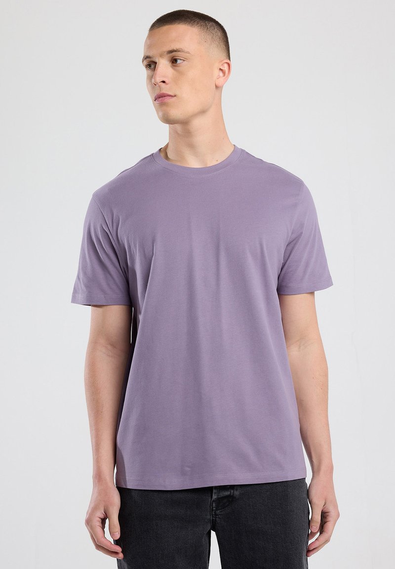 Junger Mann in einem schlichten, lavendelfarbenen T-Shirt mit kurzen Ärmeln und schwarzen Jeans, der vor einem neutralen, hellgrauen Hintergrund steht und nach links blickt.
