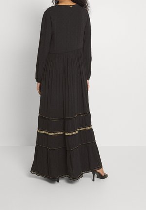 Mujer con un vestido negro largo con detalles en dorado, de pie, vista desde atrás, calzando zapatos de tacón negros.