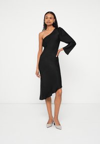 Žena v čiernych asymetrických šatách s dlhým rukávom, stojaca s rukou na páse a nosiaca béžové špicaté lodičky, na bielom pozadí.