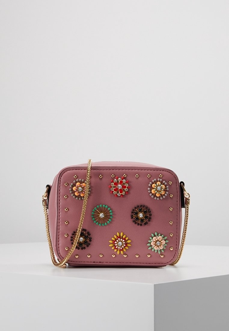 Roze leren crossbodytas met bloemrijke versieringen, gouden studs en een kettingriem. Rechthoekige vorm, gladde textuur, decoratieve accenten.
