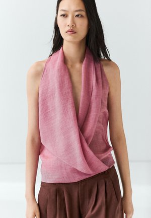 Donna con top rosa drappeggiato senza maniche e pantaloni plissettati marroni a vita alta su sfondo chiaro uniforme.