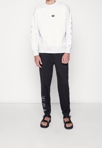 Sudadera gris claro con cuello redondo y detalle de logo, combinada con pantalones de chándal negros que presentan gráficos impresos en blanco y sandalias negras.