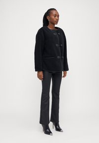 Čierna fleecová bunda s okrúhlym výstrihom, so zapínaním na kolíky a bočnými vreckami, doplnená o priliehavé čierne nohavice a špicaté podpätky.