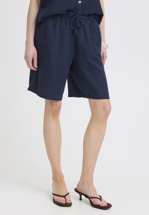 Persoană purtând pantaloni scurți bleumarin cu șnur și sandale negre cu toc, stând pe un fundal alb simplu.