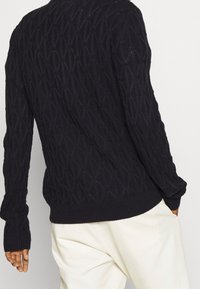 Marinblå stickad tröja med ett strukturerat mönster, ribbade ärmslut och rundad halsringning. Bakifrån framhävs en figurnära passform.