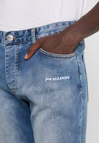 Vaqueros de mezclilla azul claro con un corte clásico, que incluyen un bolsillo frontal y el logo "PEGADOR" bordado en el muslo. Textura desgastada.
