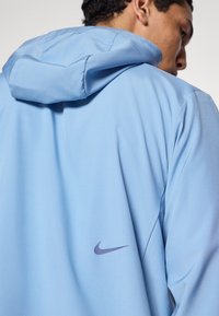 Φωτεινό γαλάζιο φούτερ Nike με κουκούλα και κορδόνι, μαλακό ύφασμα και μικρό μοβ λογότυπο στο πίσω μέρος. Λείο υφή, άνετη εφαρμογή.