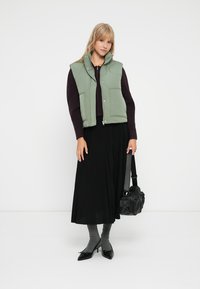 Žalias pūstas liemenė su aukštu apykakle, dėvima ant tamsios megztinio, derinama su ilgu juodu godu sijonu, pilkais kojinaitėmis, juodais kulnais ir juoda rankine.