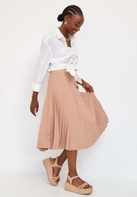 Lichtroze geplooide midi-rok, gecombineerd met een witte geknoopte blouse en beige platform sandalen met bandjes en versieringen. Soepele textuur.