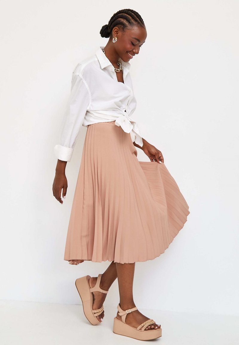 Lichtroze geplooide midi-rok, gecombineerd met een witte geknoopte blouse en beige platform sandalen met bandjes en versieringen. Soepele textuur.